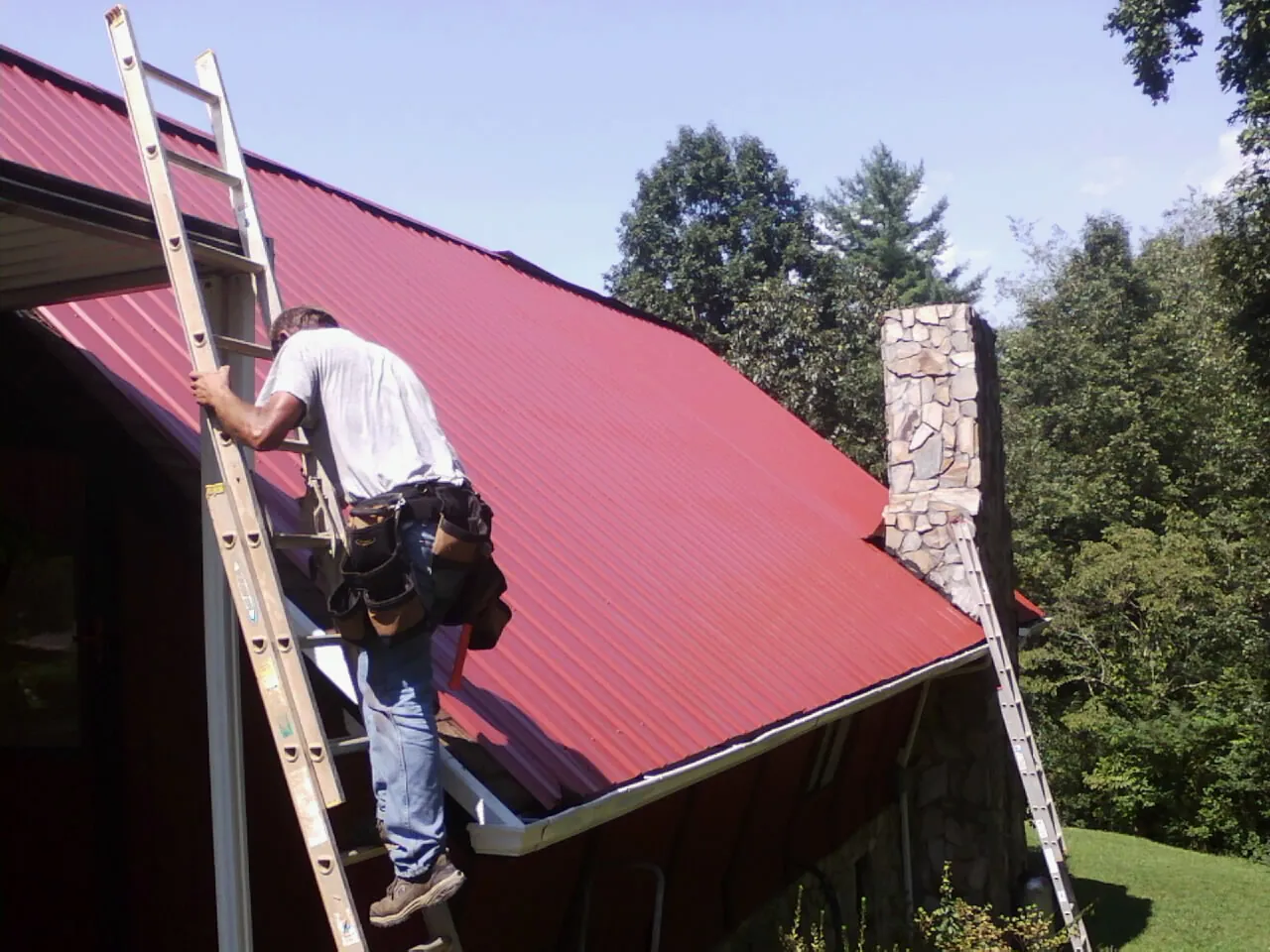 Roofer on a ladder beside a red metal roof and stone chimney — Soffit & Fascia Repair by Roofing Brothers in Kanab